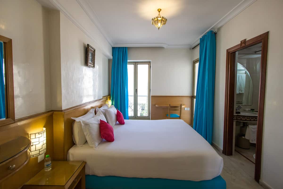Une chambre d’hôtel avec un grand lit, une tête de lit en bois, une table de chevet avec une lampe, un miroir et une vue sur l’extérieur par une fenêtre ornée de rideaux bleus.