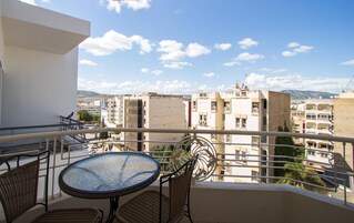 Uma varanda com cadeiras de vime e uma mesa de vidro, com vista para um horizonte urbano com edifícios e montanhas.