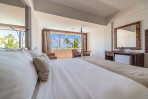 A hotel room with a large bed, a desk, a chair, a small table, a mirror, and a view of the beach.