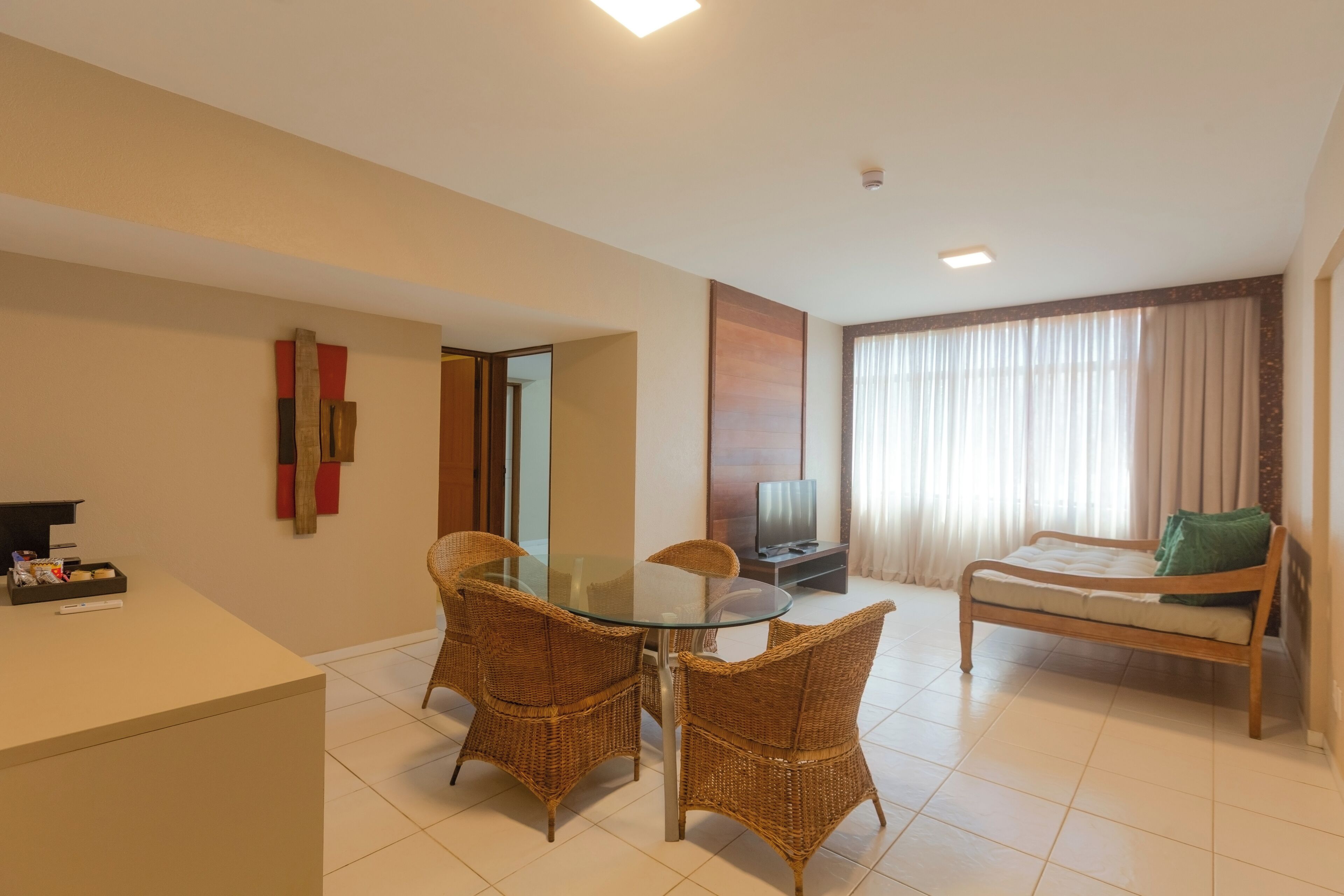 A hotel room with a glass-top dining table, wicker chairs, a bed with a green pillow, a TV, and a reception desk.