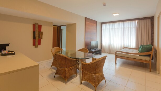 A hotel room with a glass-top dining table, wicker chairs, a bed with a green pillow, a TV, and a reception desk. - Ritz Lagoa Da Anta (Maceió)