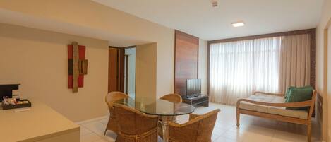 A hotel room with a glass-top dining table, wicker chairs, a bed with a green pillow, a TV, and a reception desk.