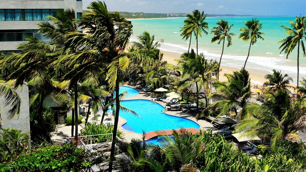 Een resort met een zwembad, omringd door palmbomen, een strand en een modern gebouw.