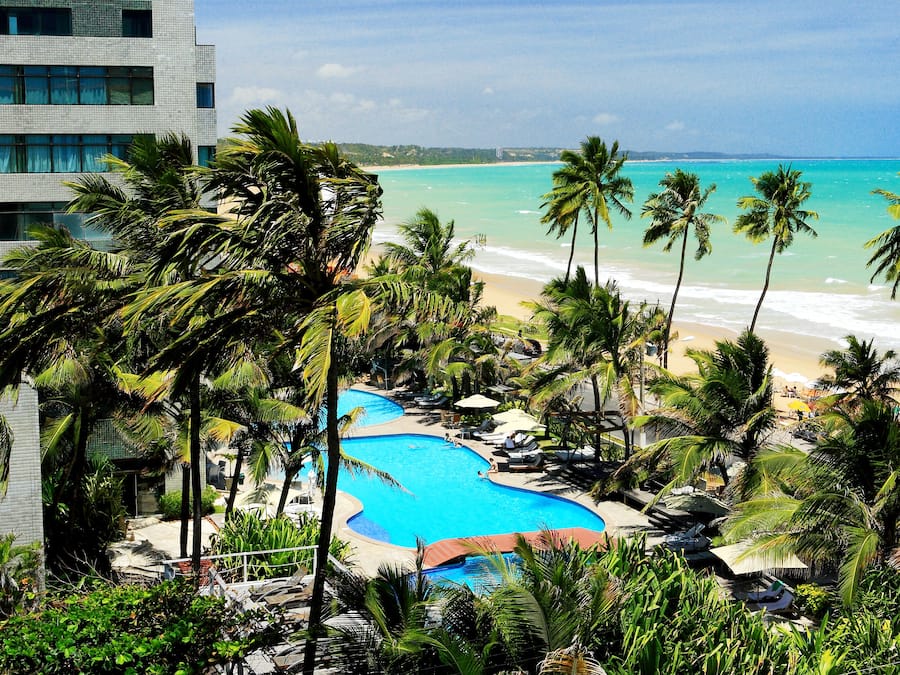 Un resort con piscina rodeada de palmeras, playa y un edificio moderno.