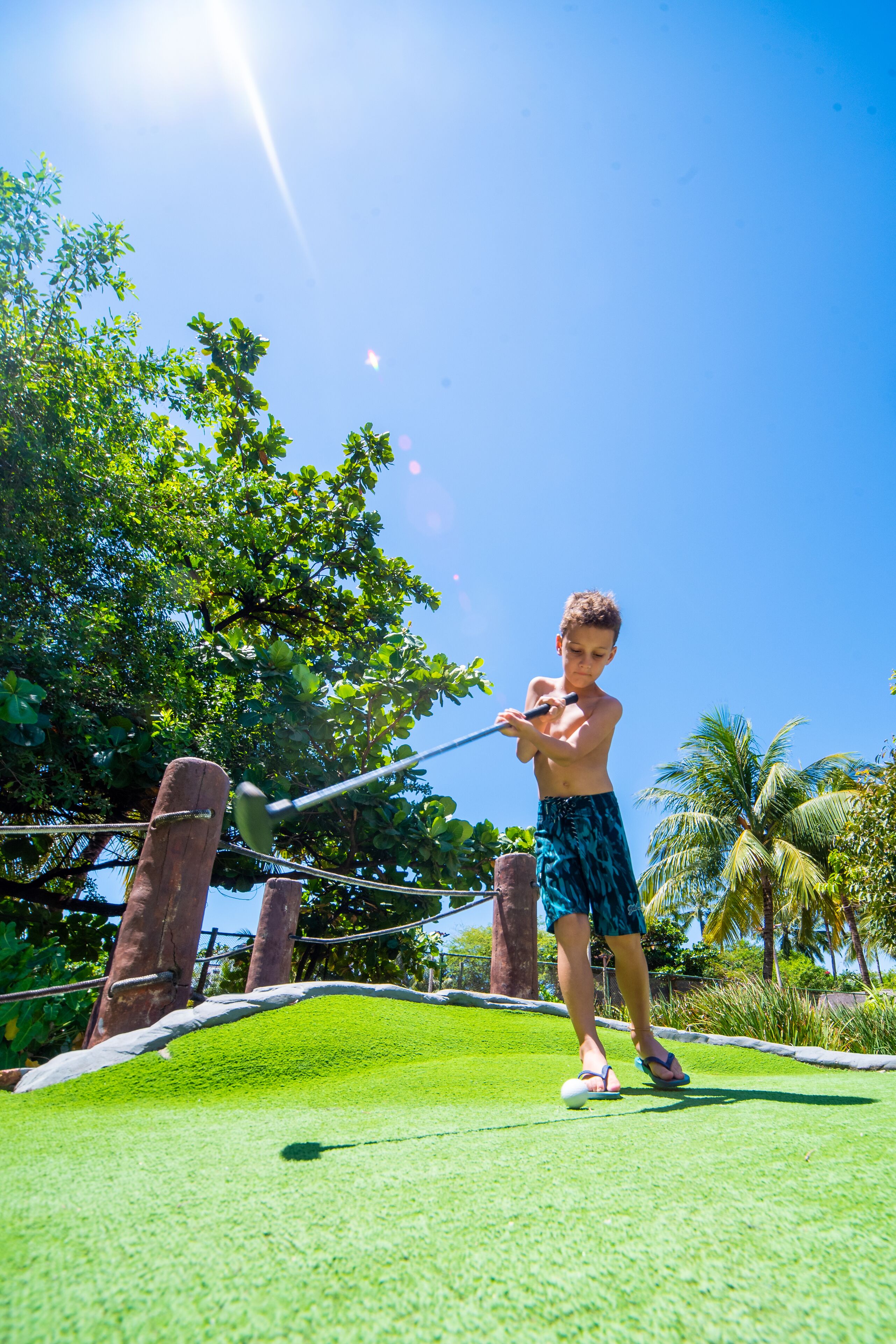A person playing golf on a green field with trees and palm trees in the background.