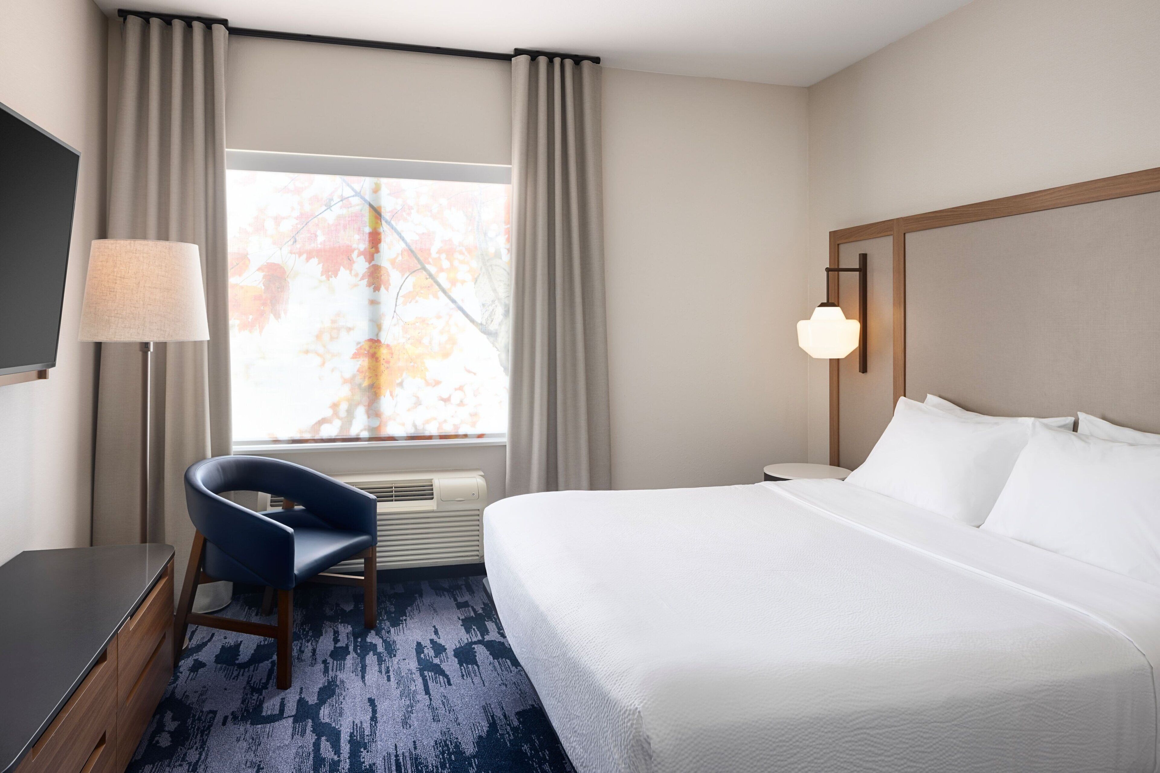 Une chambre d’hôtel avec un lit, un bureau, une chaise, une lampe et une fenêtre avec des rideaux.