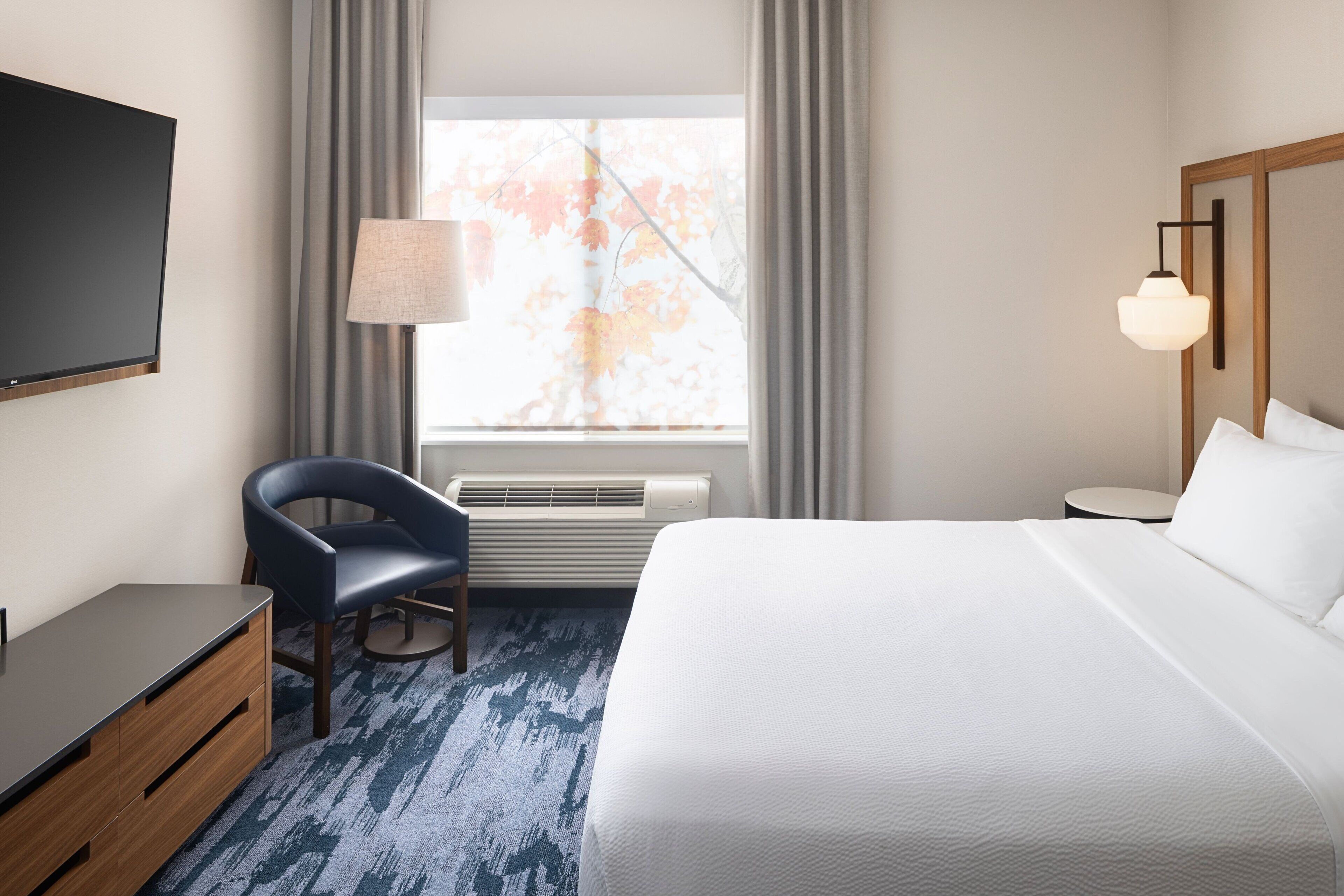 Une chambre d’hôtel avec un lit, un bureau, une chaise, une télévision, une lampe et une fenêtre avec des rideaux.