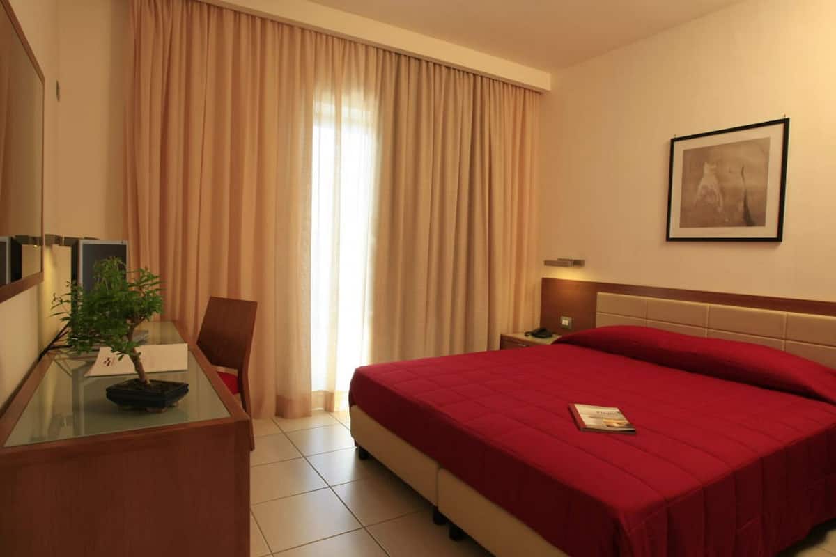 A hotel room with a bed, a desk with a bonsai tree, a chair, a nightstand, and a framed picture on the wall.