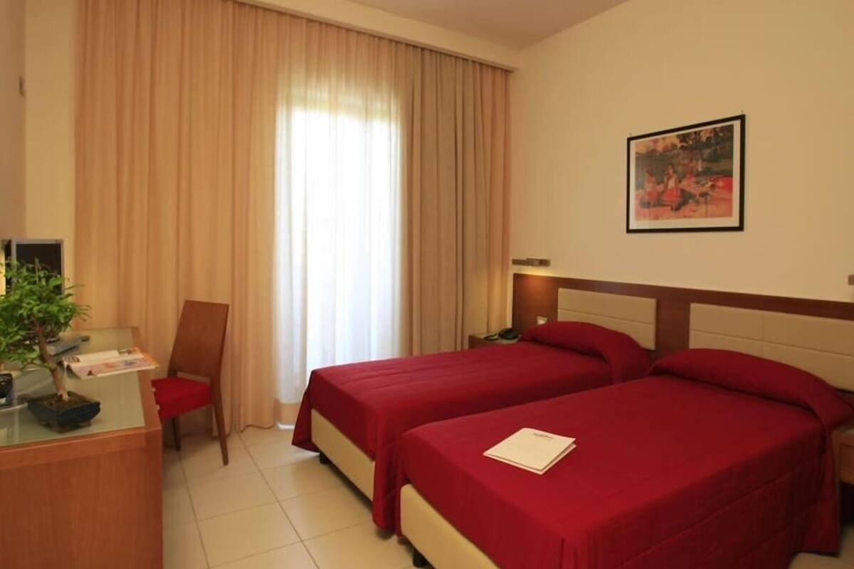 A hotel room with two beds, a desk, a chair, a window with curtains, and a framed picture on the wall.