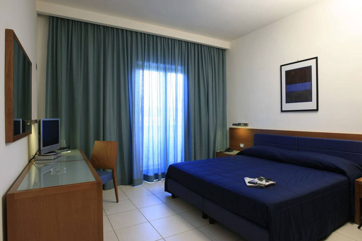 A hotel room with a bed, a desk with a computer, a chair, a nightstand, and a window with curtains.