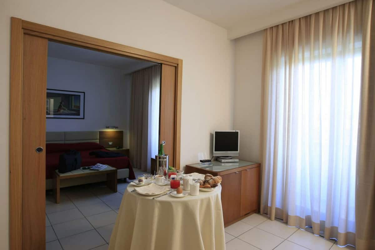 A hotel room with a dining table set for breakfast, a TV on a stand, a sofa, and a painting on the wall.