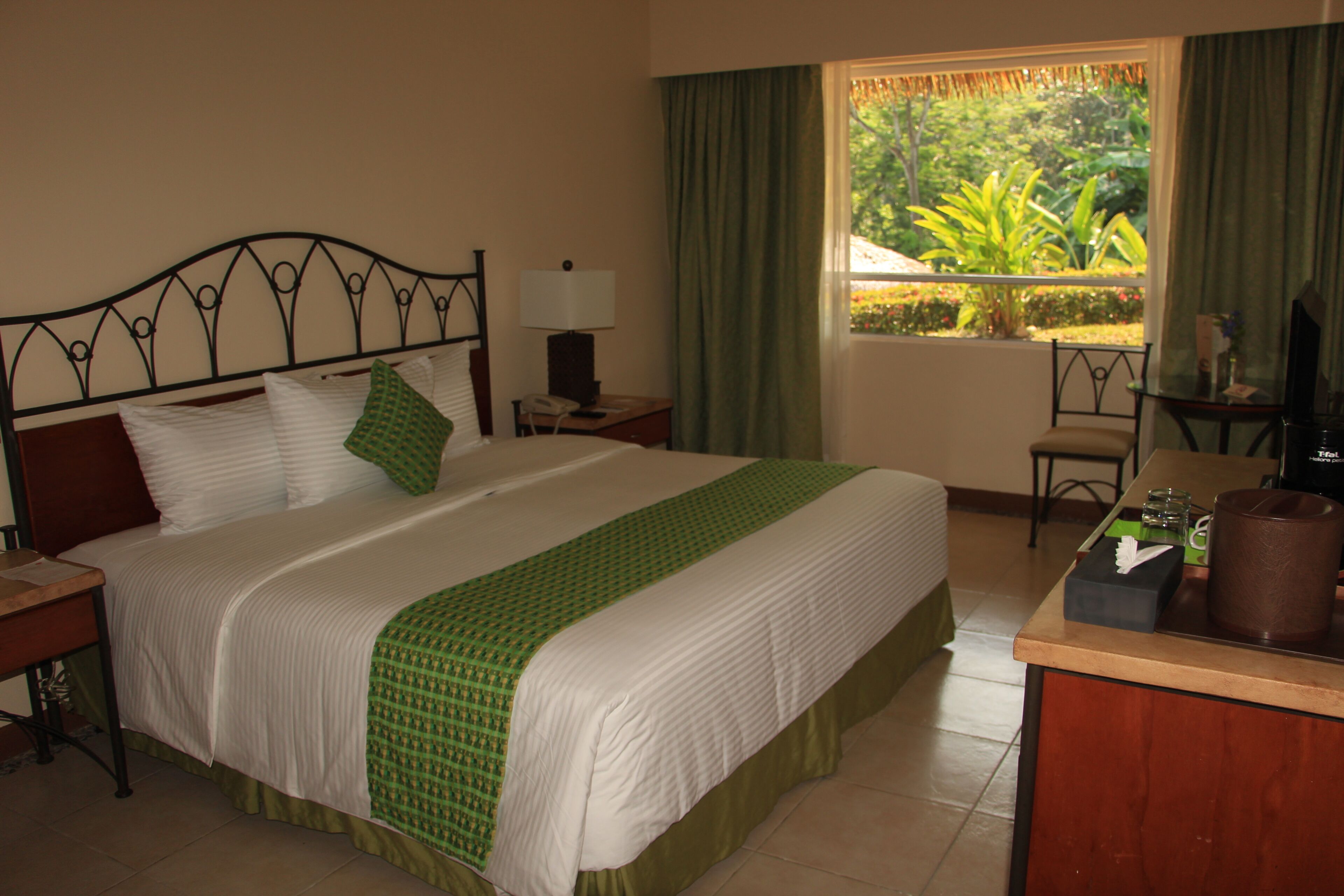A neatly made bed with a green and white bedspread, a bedside table with a lamp, a chair, and a view of greenery outside the window.