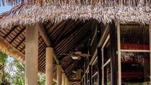 A covered outdoor area with a thatched roof, wooden tables, chairs, and a pathway leading to a building.