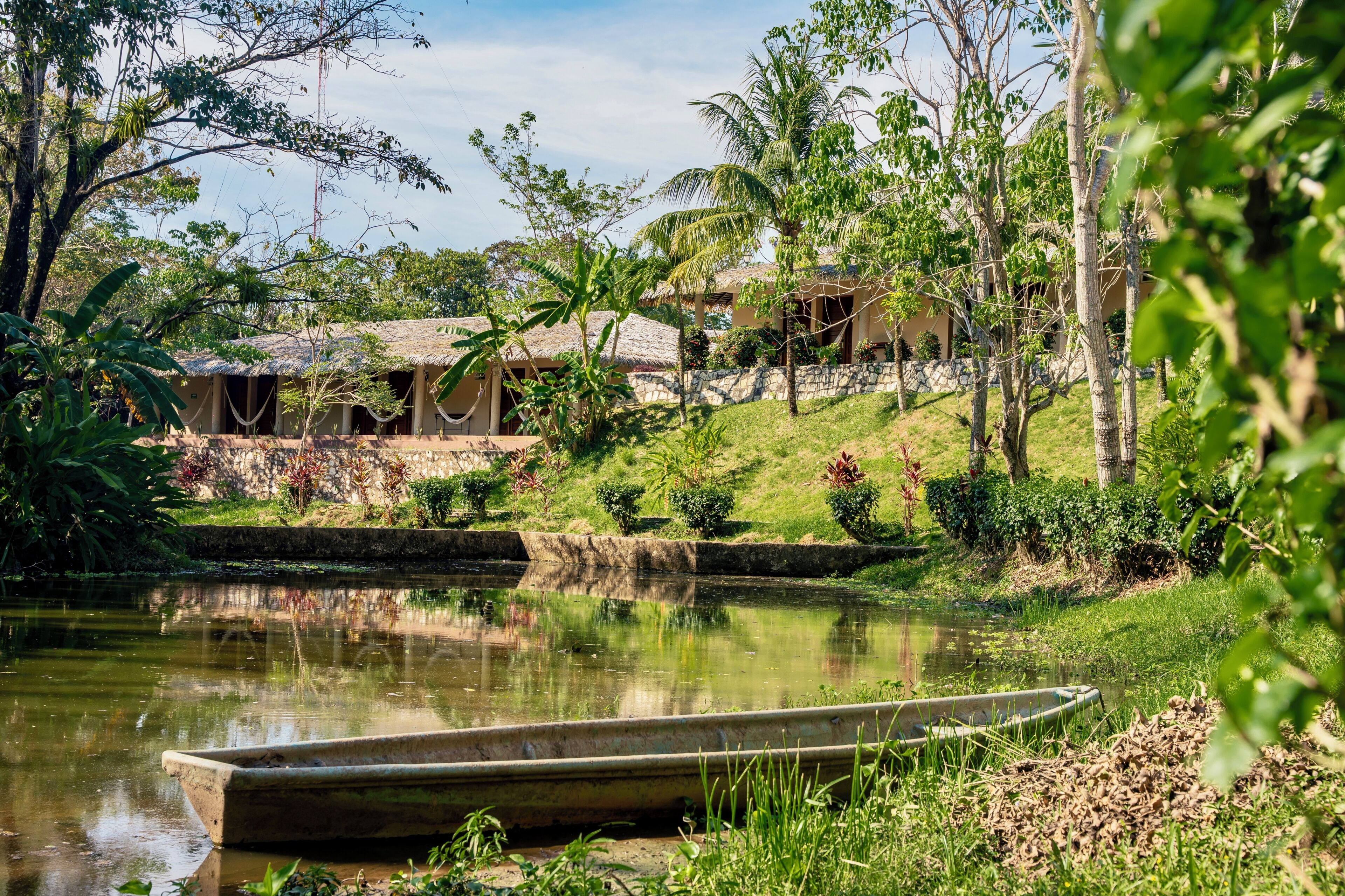 A calm pond with a boat, surrounded by lush greenery and a building with a thatched roof.