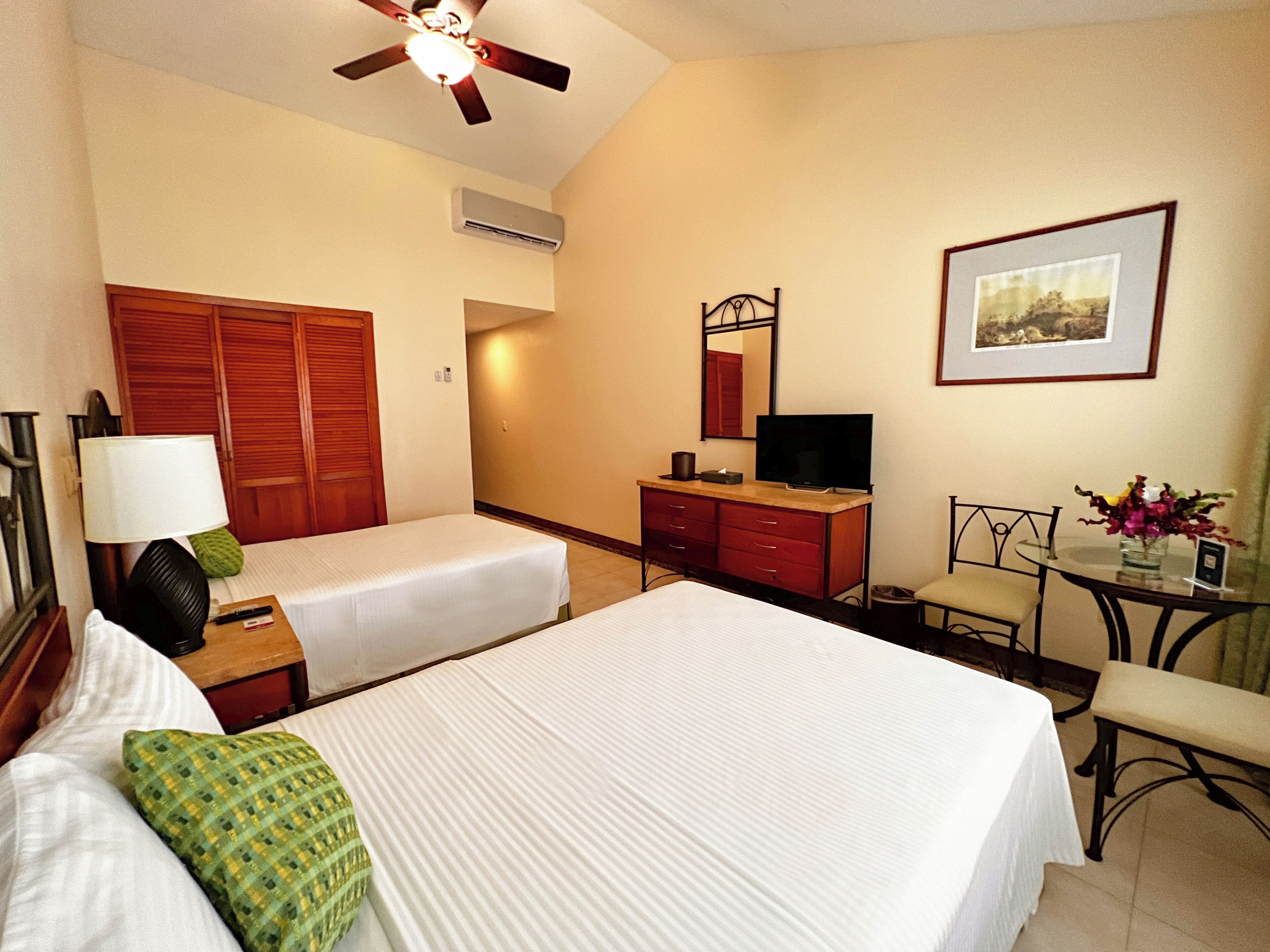 A hotel room with two beds, a desk, a television, a mirror, and a ceiling fan.