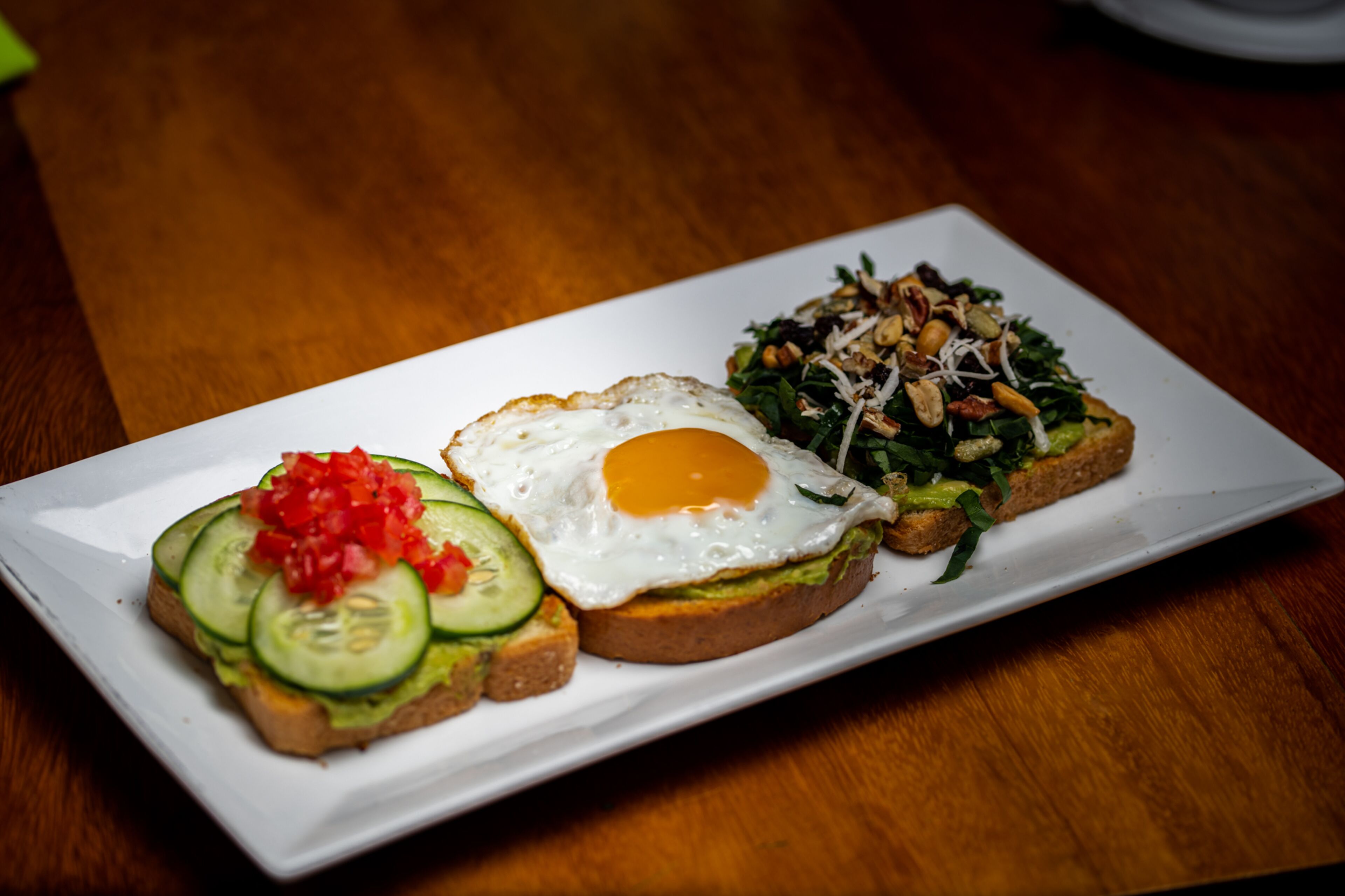 A white rectangular plate with three different food items: a fried egg, a sandwich with cucumber slices, and a salad with nuts.