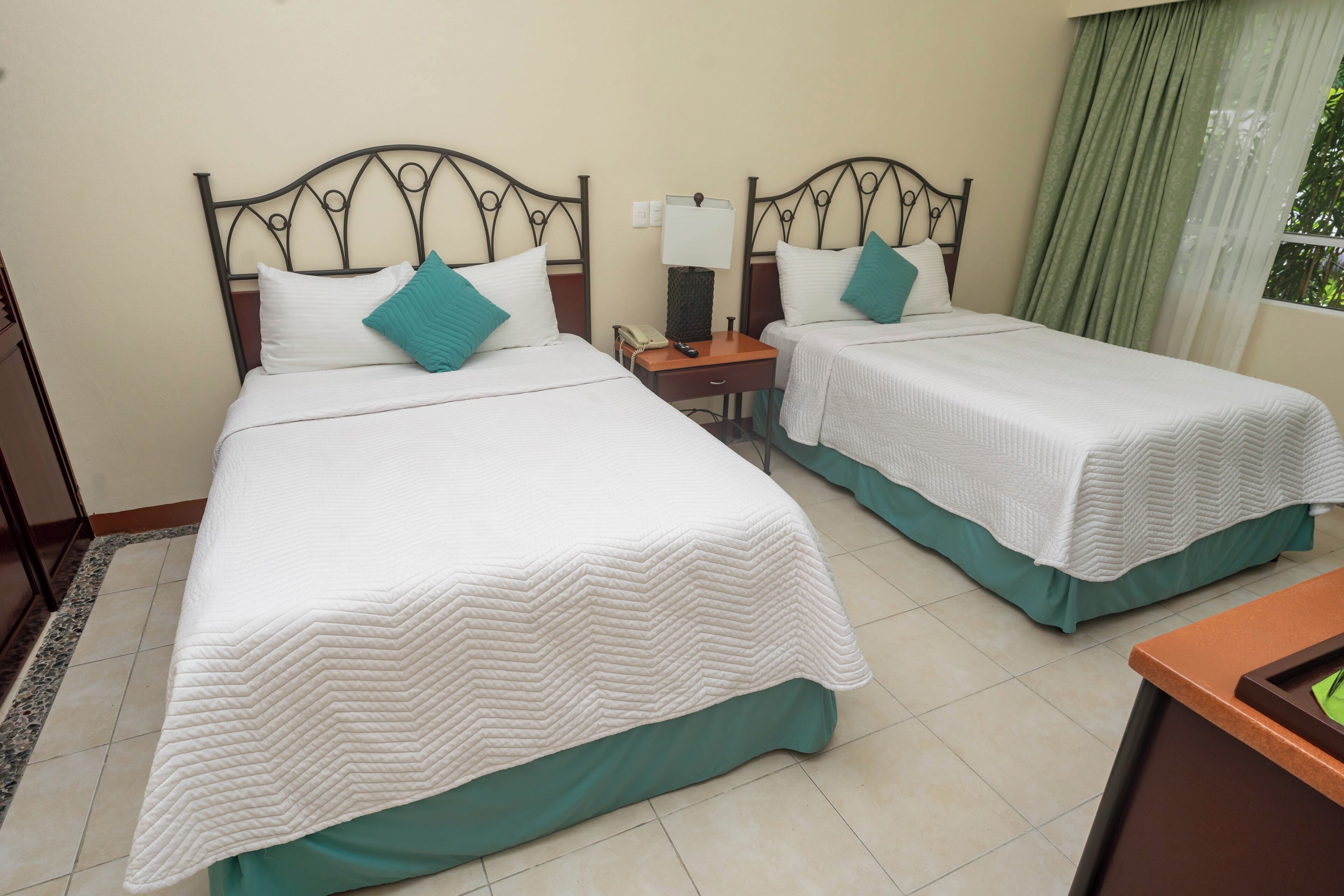 Habitación de hotel con dos camas, cada una con cubrecama blanco y almohadas turquesa, una mesita de noche con lámpara y una ventana con cortinas verdes.
