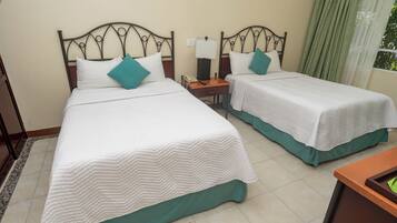 Habitación de hotel con dos camas, cada una con cubrecama blanco y almohadas turquesa, una mesita de noche con lámpara y una ventana con cortinas verdes.