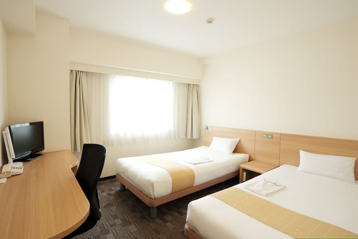 Una habitación de hotel con dos camas, un escritorio con televisor y una ventana con cortinas.