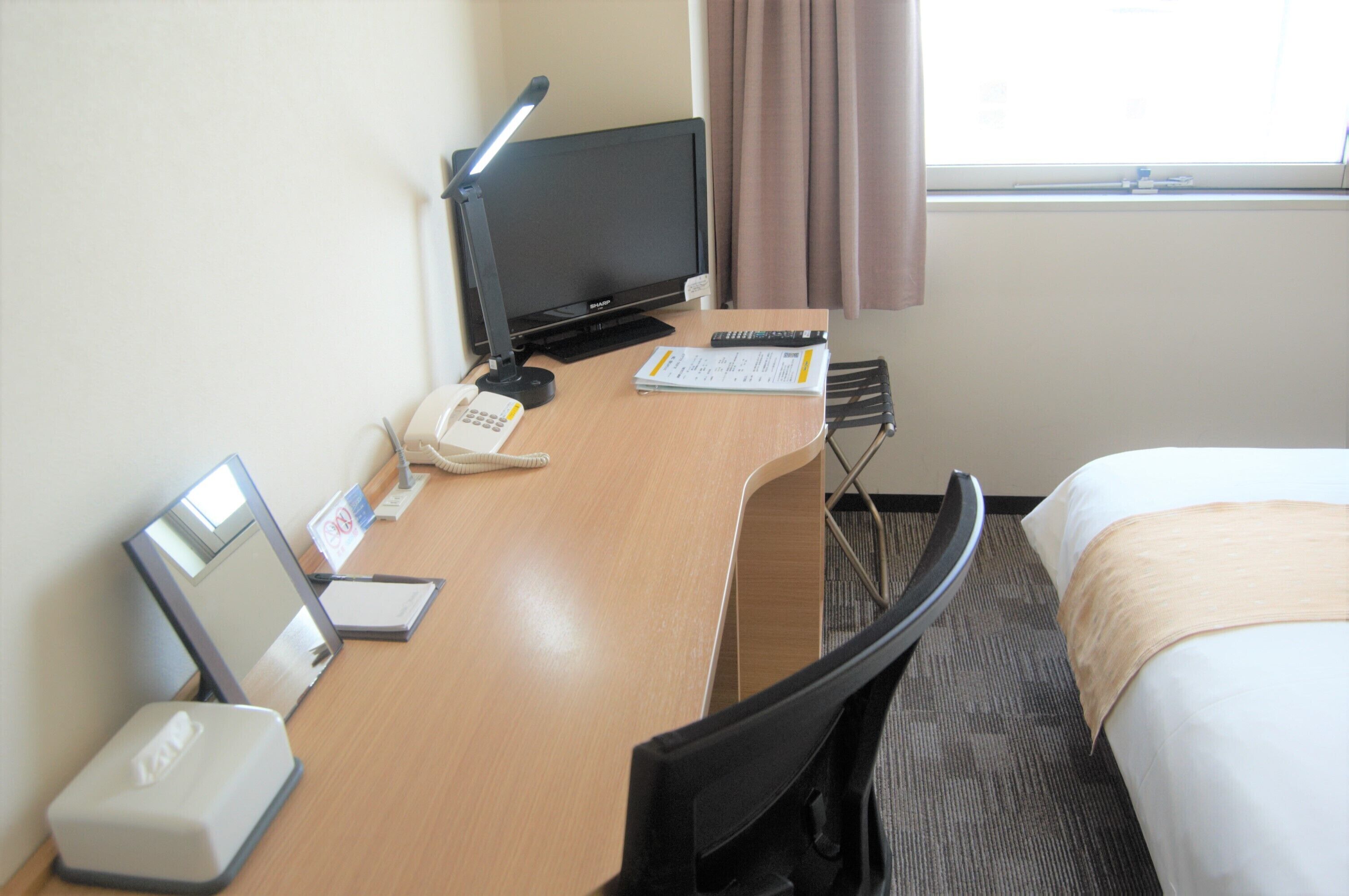 Habitación de hotel con escritorio, computadora, teléfono, lámpara, cama y ventana con cortinas.