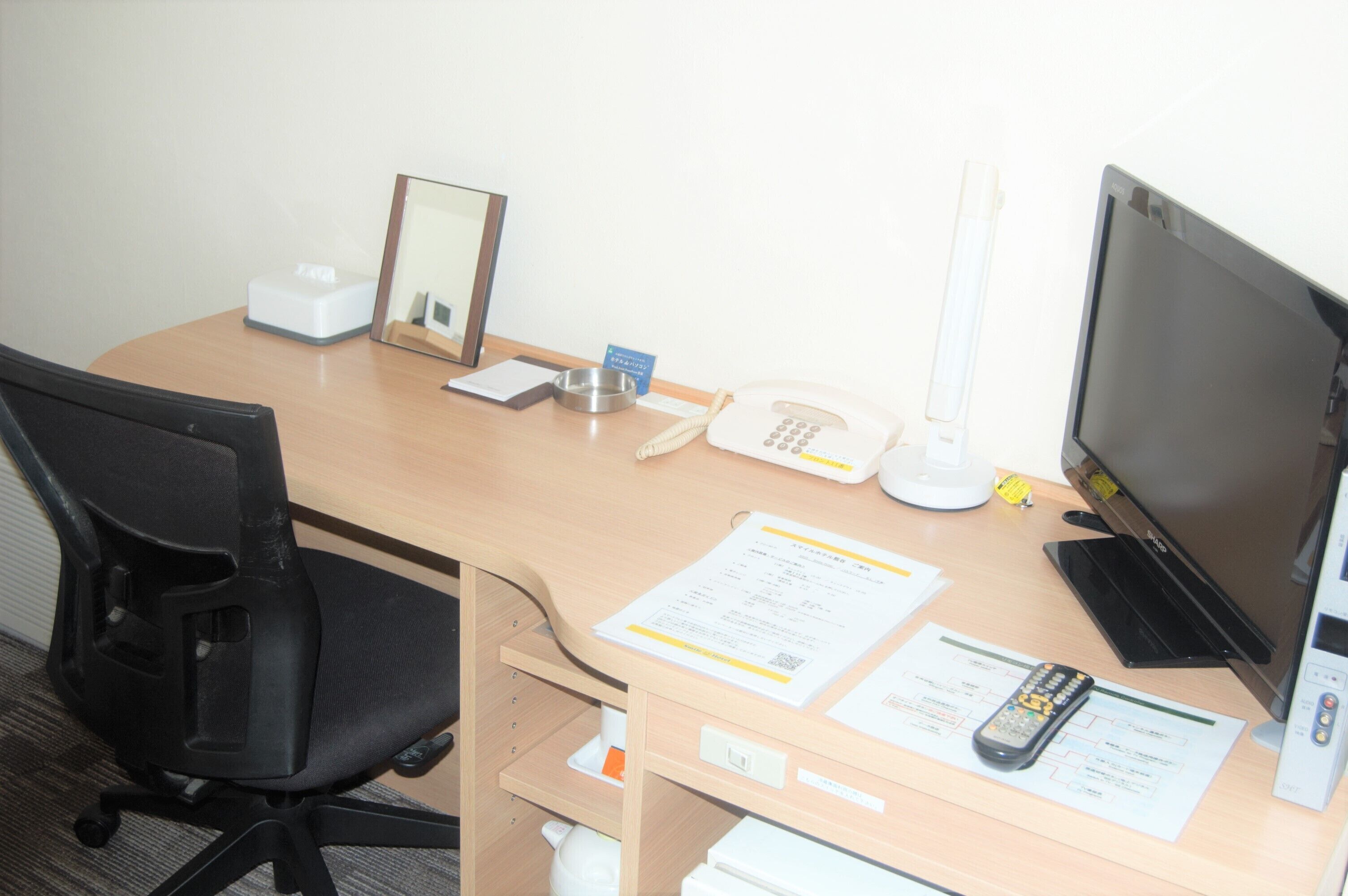 A hotel room with a wooden desk, a black office chair, a telephone, a remote control, and a television.