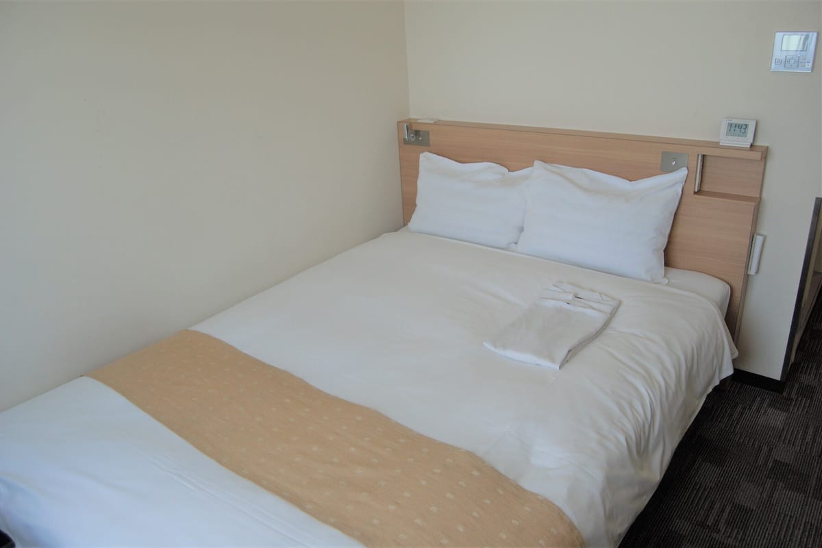 Una cama bien hecha con ropa de cama blanca y beige, un cabecero de madera y un reloj digital en la pared.