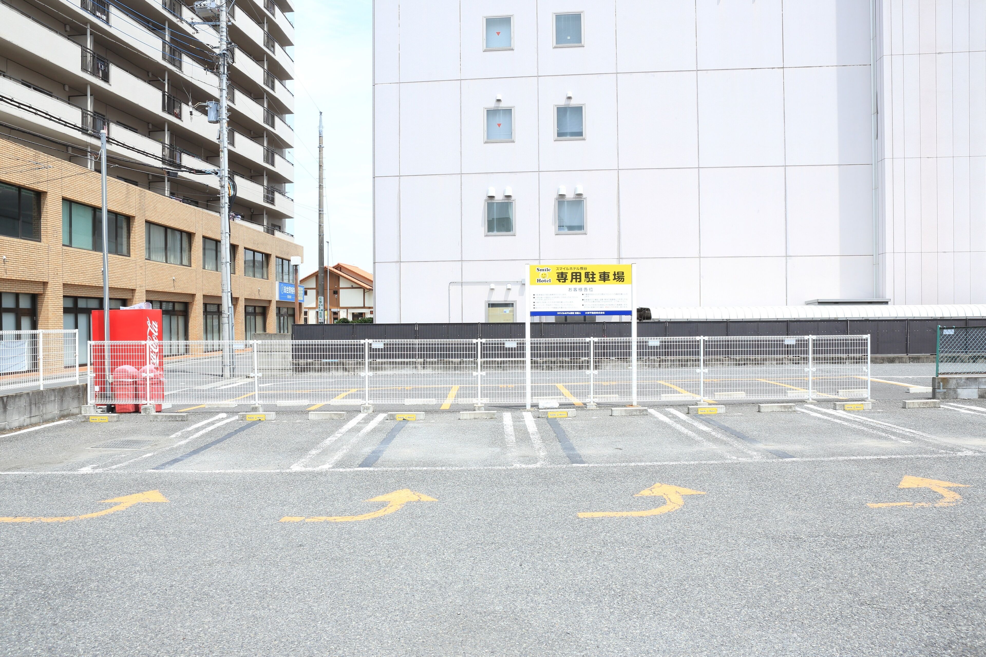 A parking lot with directional arrows, a fenced area, and a building with multiple floors in the background.