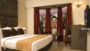 A hotel room with a large bed, a desk with a chair, a television, and a view of the city at dusk. - Shilton Royale (Bengaluru)