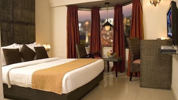 A hotel room with a large bed, a desk with a chair, a television, and a view of the city at dusk.