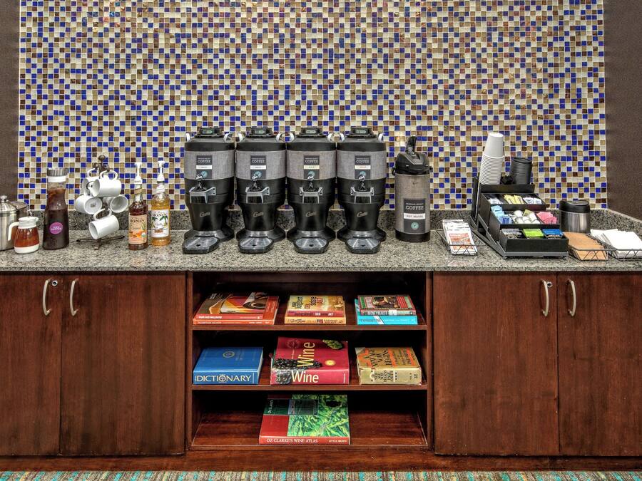 A coffee station with multiple coffee makers and a coffee machine, a selection of coffee pods, and a coffee grinder. There are also several books and a wine bottle on the counter.