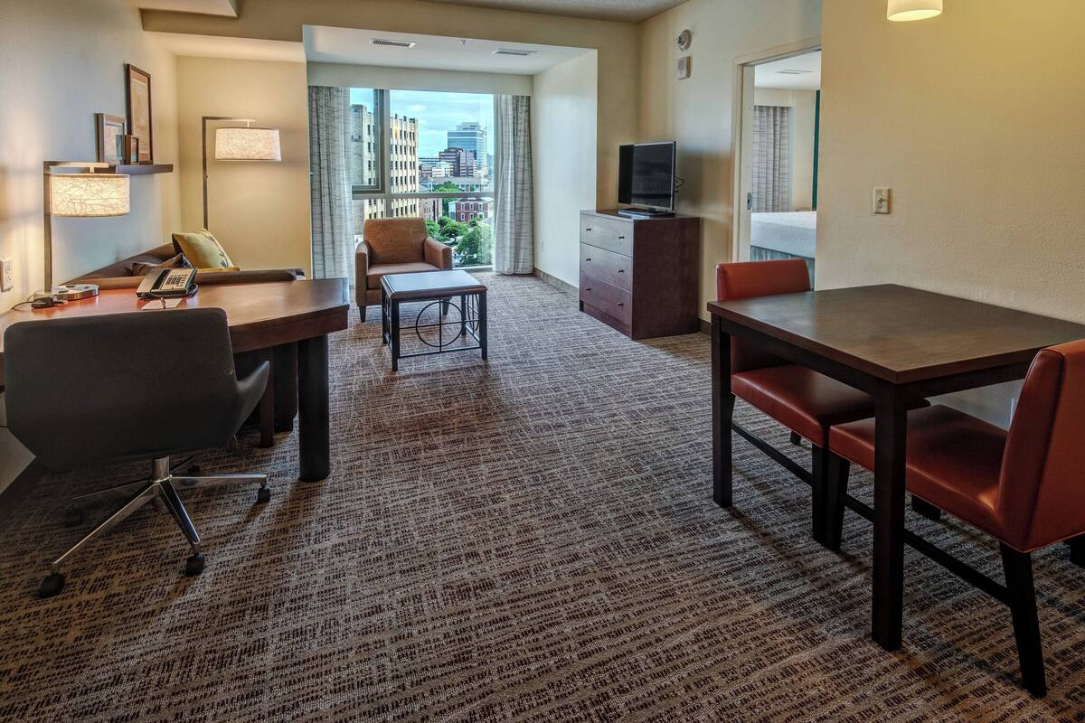 A hotel room with a large window, a desk with a chair, a dining table with chairs, a television on a dresser, and a carpeted floor.