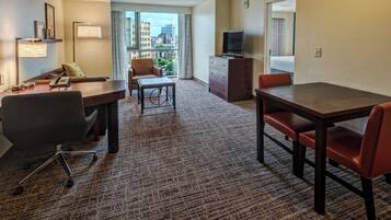 A hotel room with a large window, a desk with a chair, a dining table with chairs, a television on a dresser, and a carpeted floor.