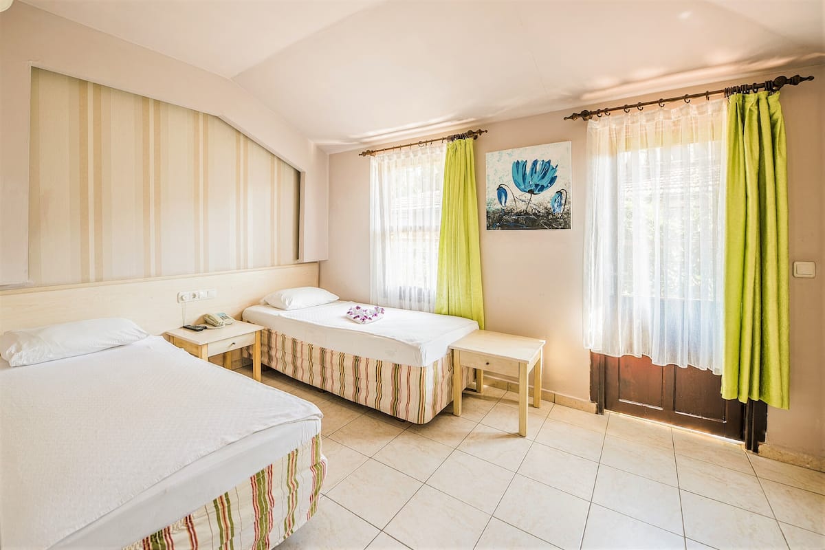 Hotellihuone, jossa on kaksi sänkyä, yöpöytä, ikkuna verhoilla ja seinällä taulu.