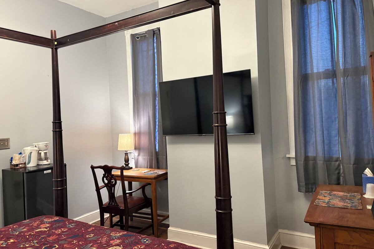 A bedroom with a four-poster bed, a desk with a chair, a television mounted on the wall, and a window with curtains.