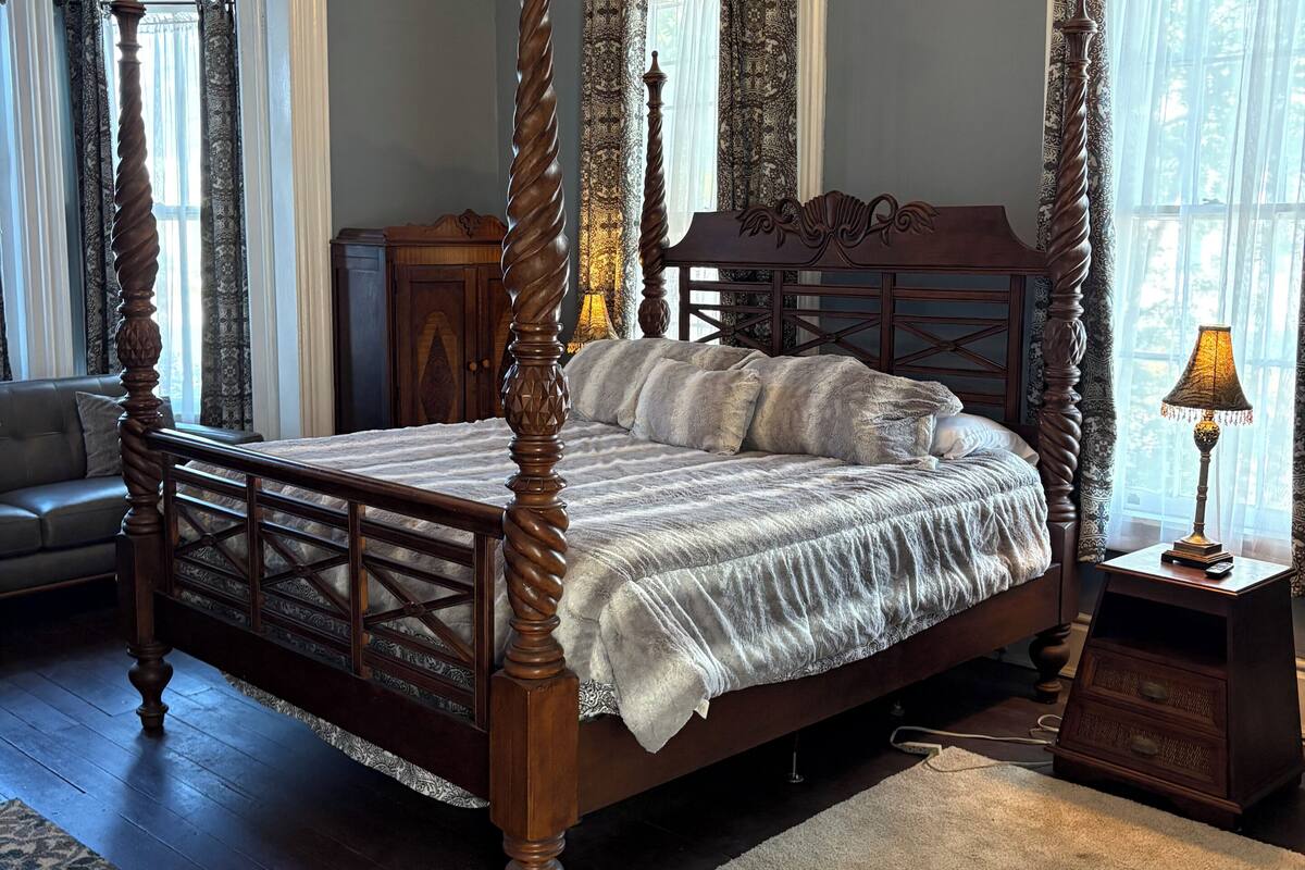 A four-poster bed with a wooden frame and a white bedspread, a chandelier, and a nightstand with a lamp.