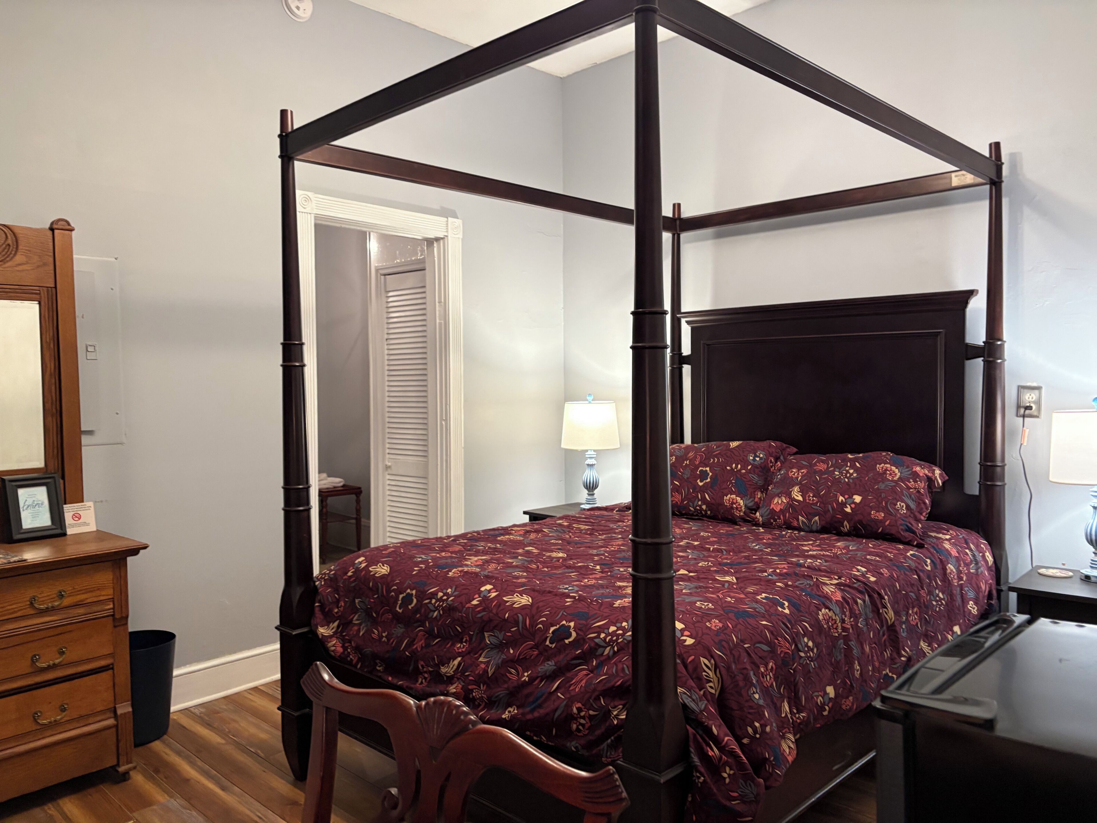 Een hemelbed met een bloemenprint, een houten dressoir, een nachtkastje met een lamp en een kast met een witte deur.