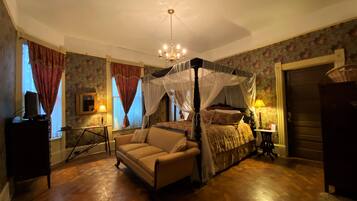 A bedroom with a four-poster bed, a sitting area with a sofa, a wooden floor, and a chandelier.