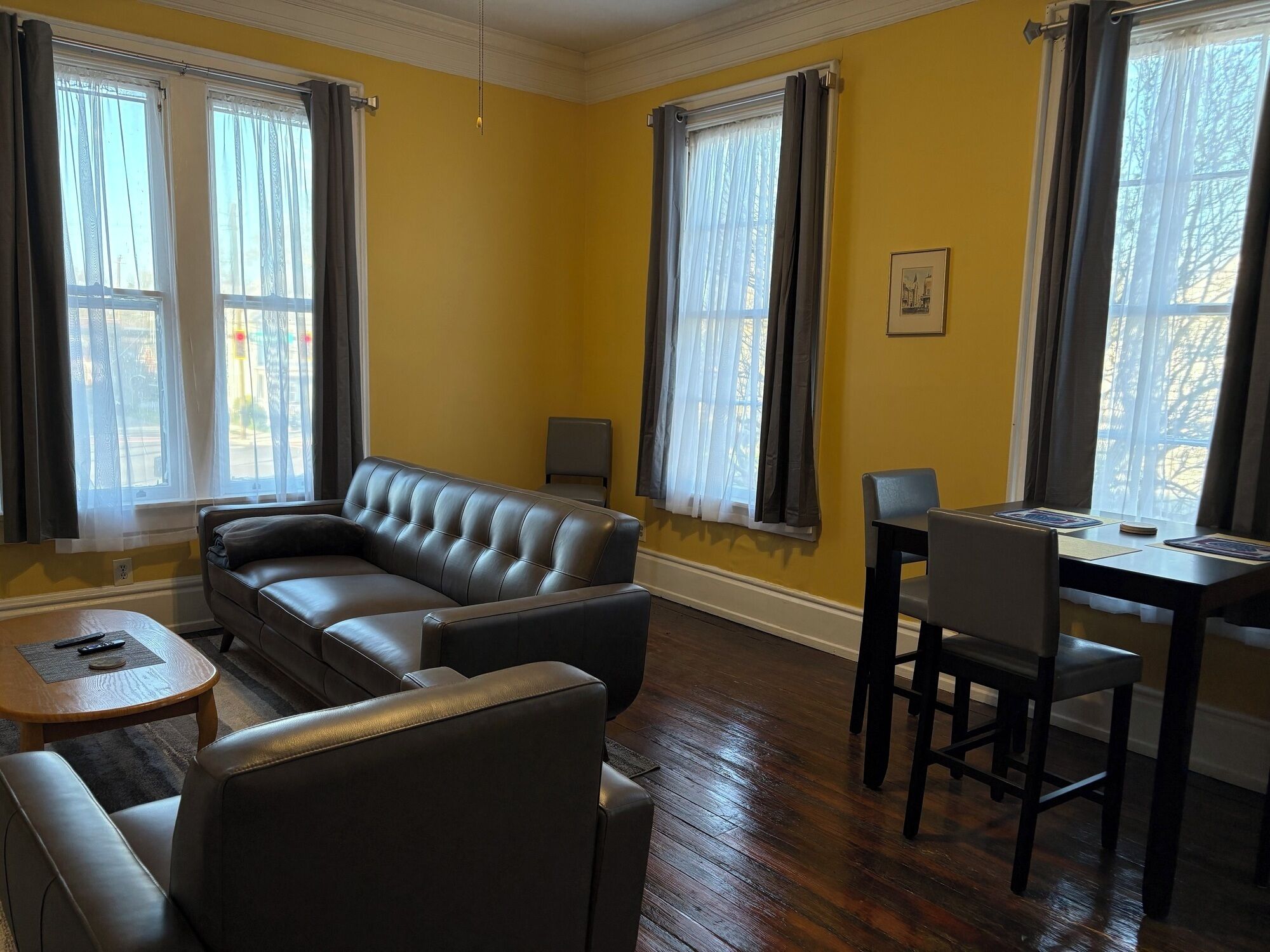A living room with a sofa, a small table, a chair, and a dining table with chairs. There are windows with curtains and a framed picture on the wall.