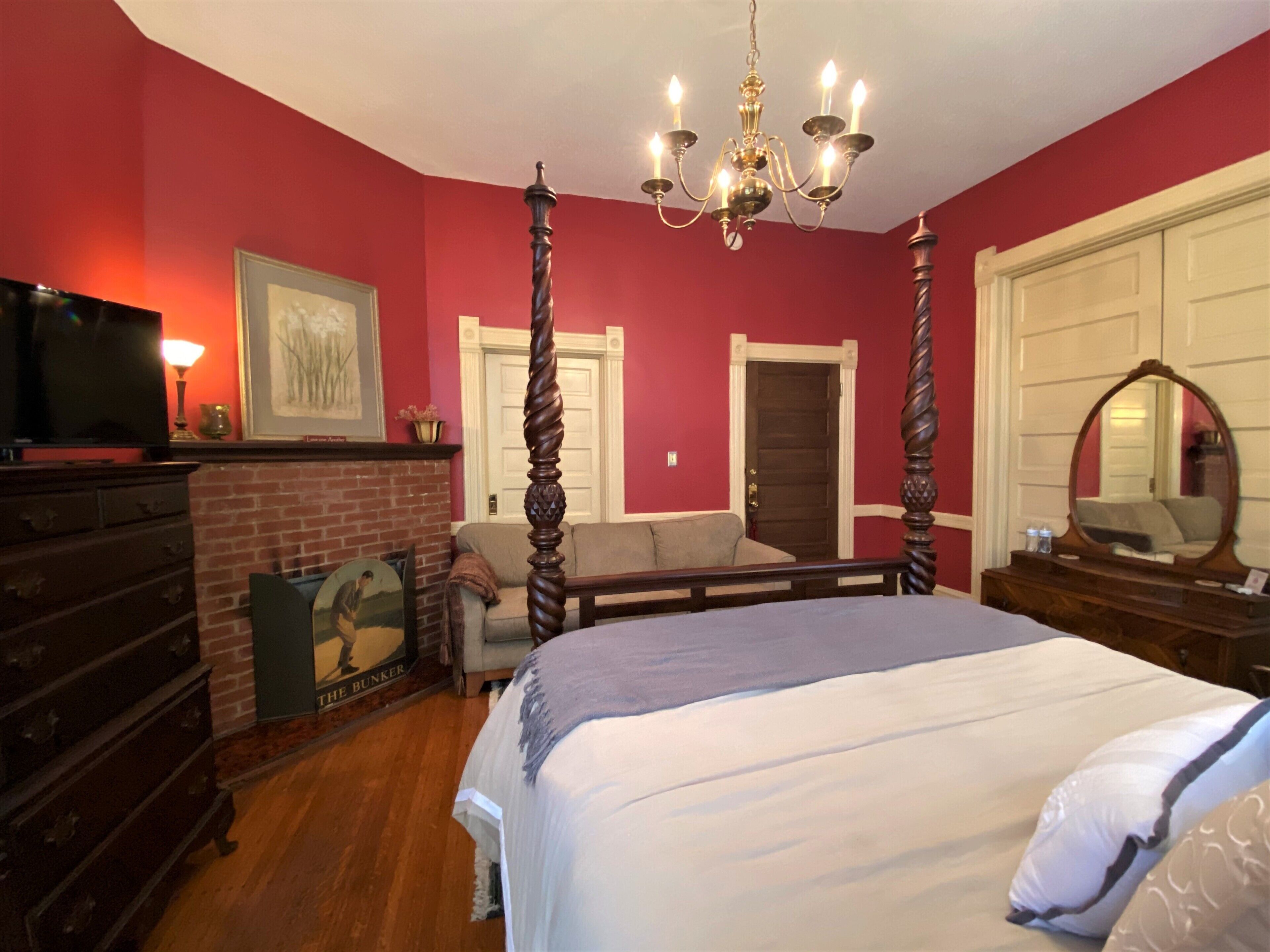 A bedroom with a four-poster bed, a chandelier, a television on a dresser, and a fireplace.