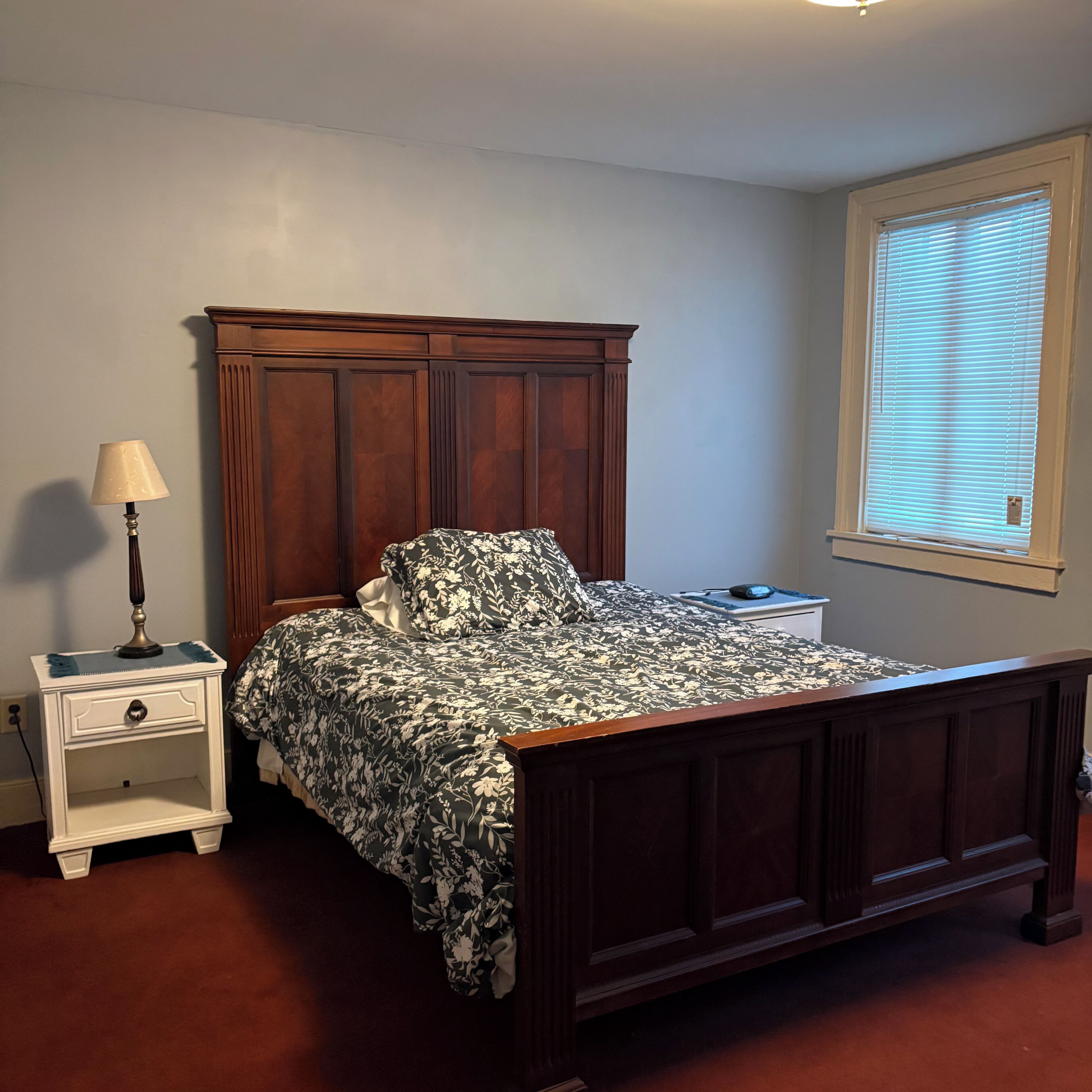 A bedroom with a wooden bed, a nightstand, a lamp, and a window with blinds.