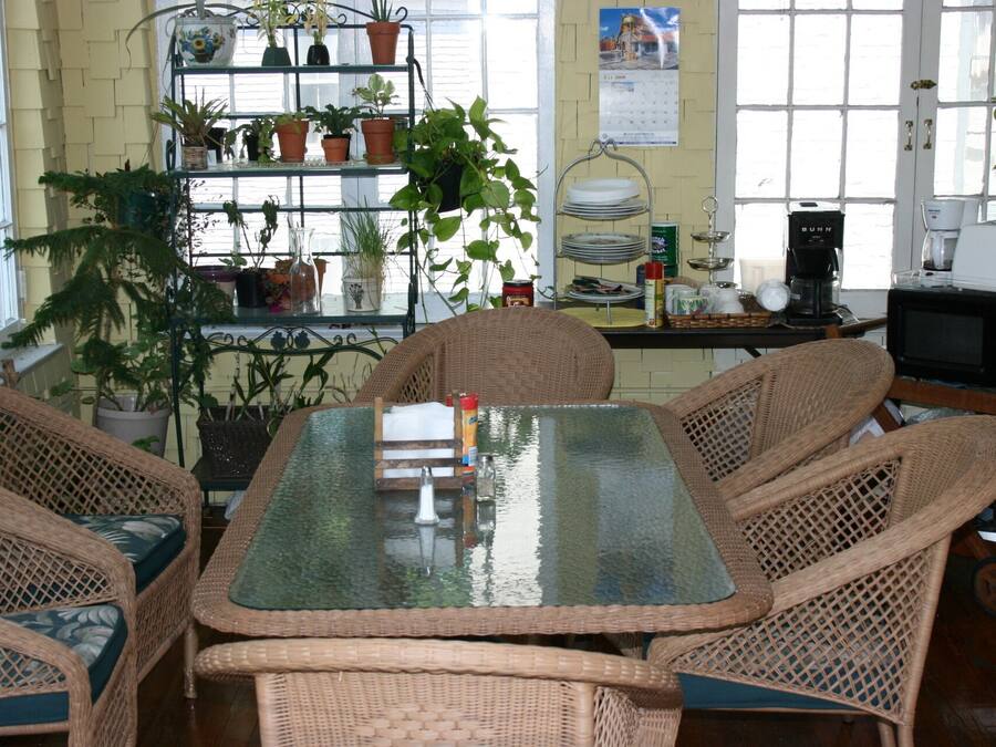 A dining area with wicker chairs and a glass table, surrounded by potted plants and kitchen appliances.