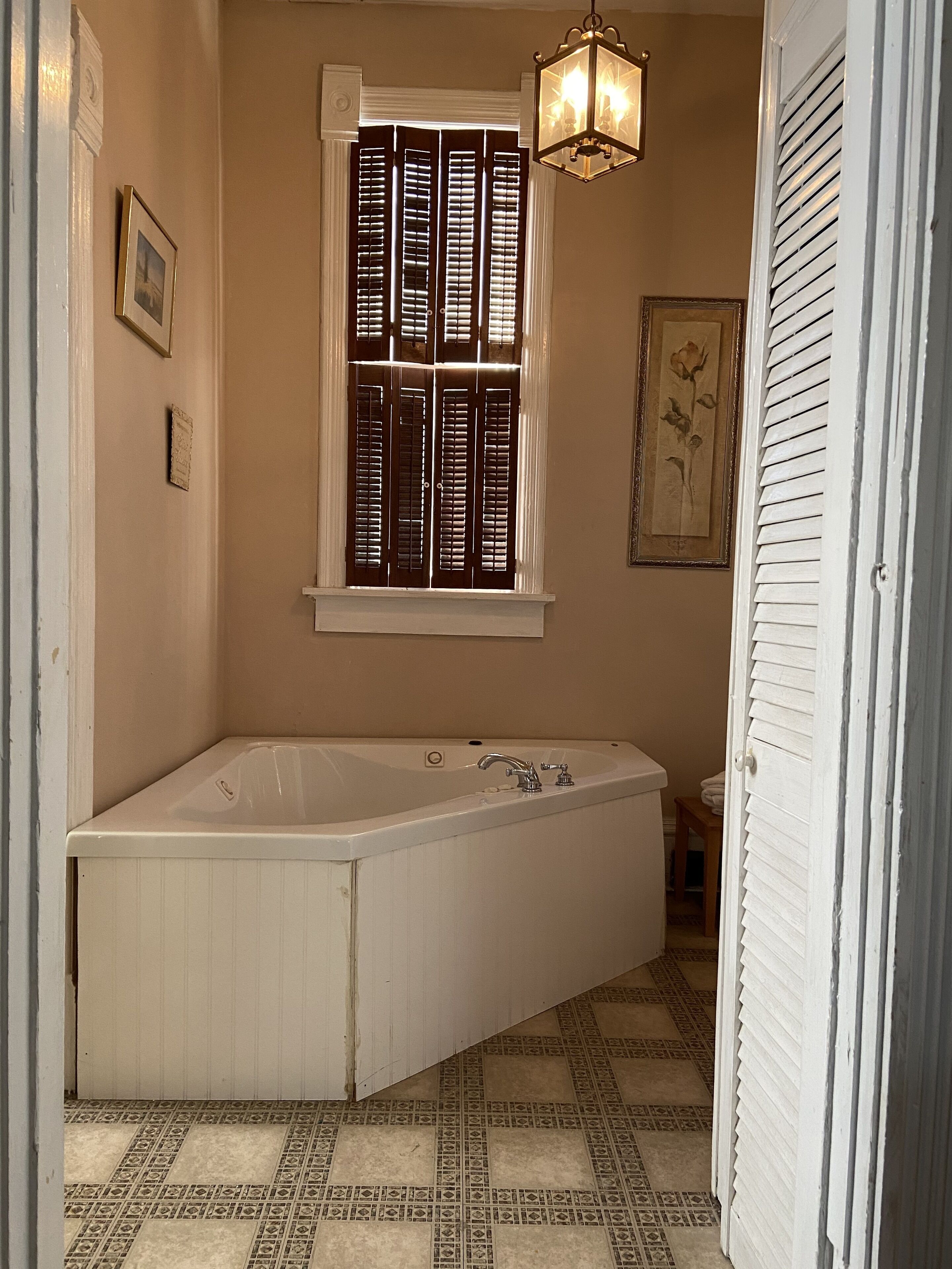 A bathroom with a freestanding bathtub, a window with shutters, a hanging light fixture, and patterned floor tiles.