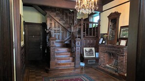 A grand staircase with intricate woodwork, a chandelier, and a fireplace with a brick surround. - The Queen Anne Inn (Augusta)