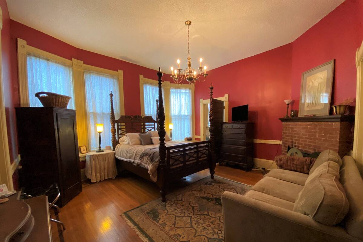A bedroom with a four-poster bed, a chandelier, a fireplace, a sofa, and a dresser.