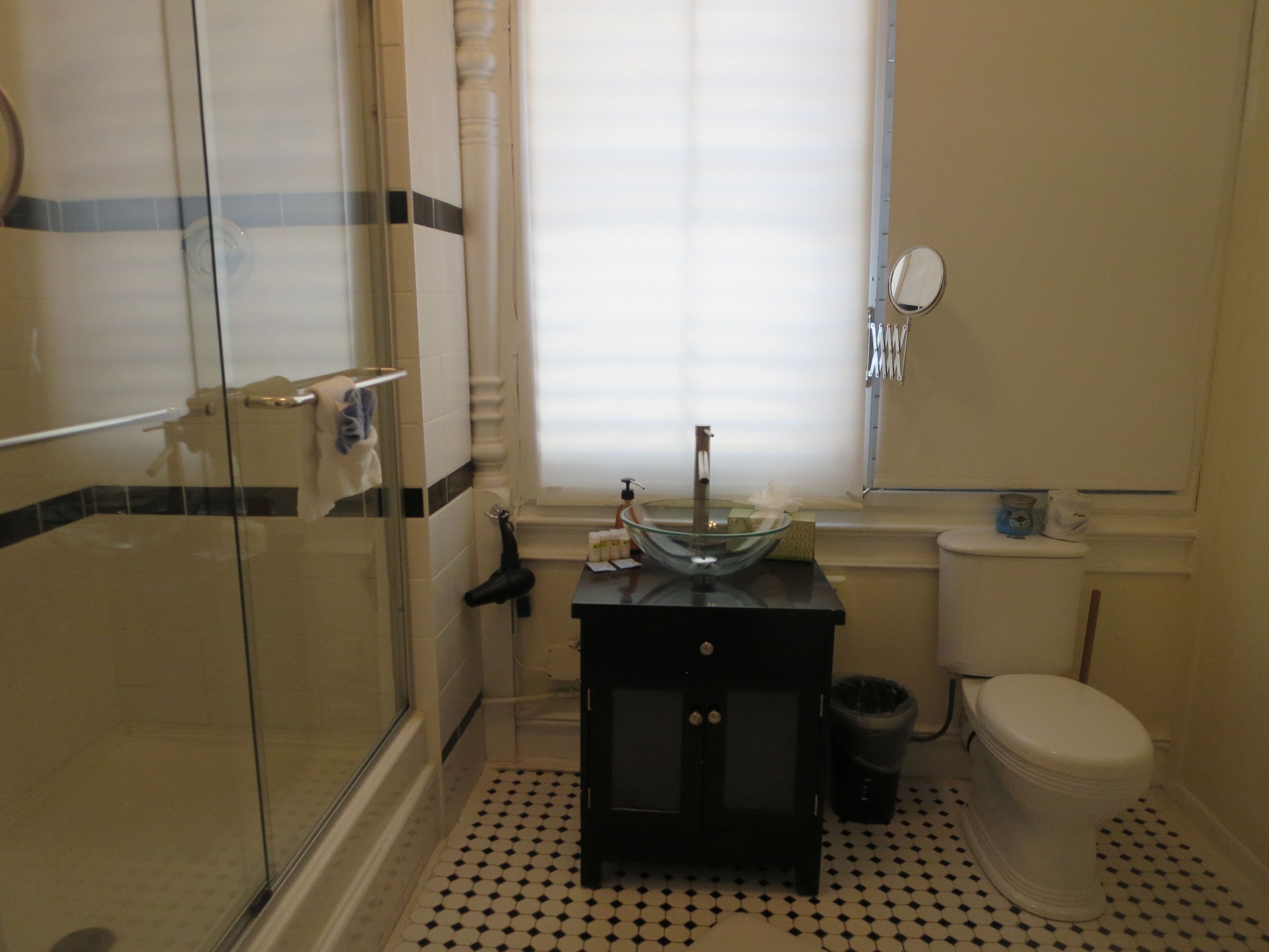 A bathroom with a glass shower enclosure, a black sink with a glass basin, a toilet, and a window with blinds.