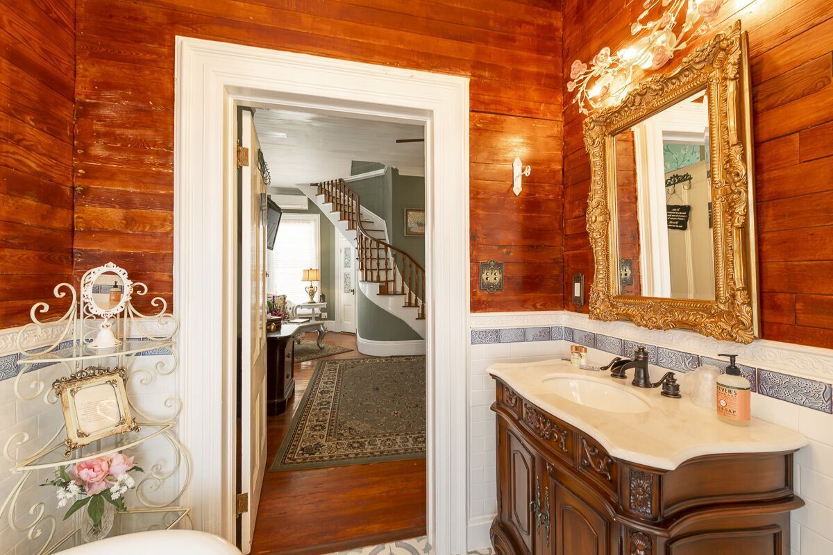 A bathroom with a wooden wall, a large ornate mirror, a sink with a soap dispenser, and a decorative metal rack with a framed picture and flowers.