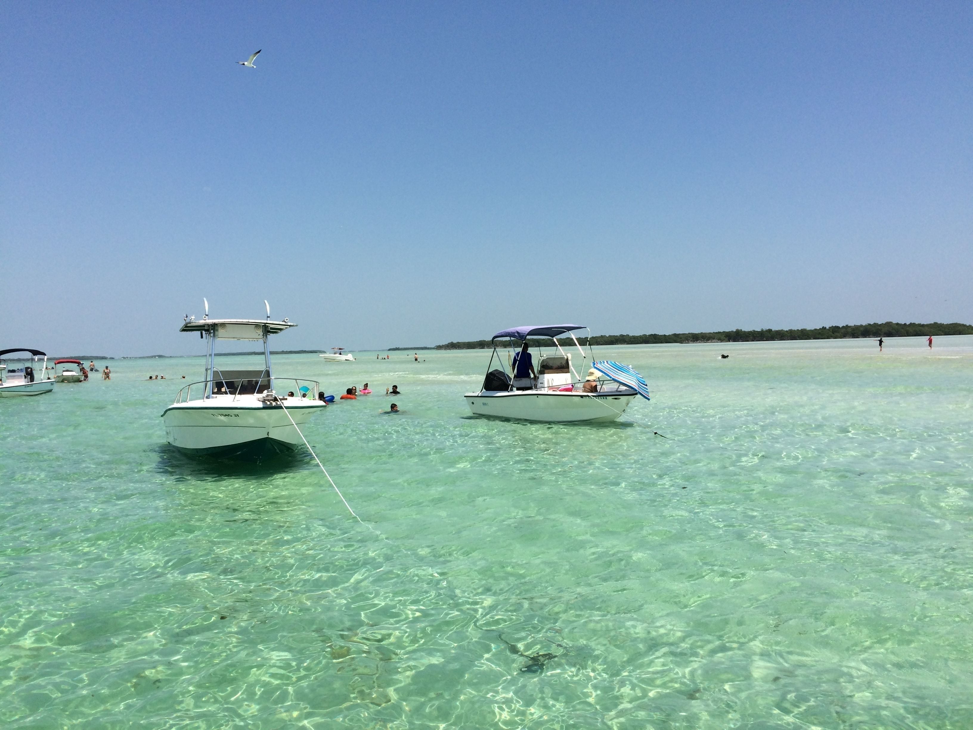 Un mar transparente y poco profundo con barcos fondeados, una playa de arena y un cielo despejado.
