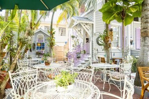 Un área de patio con mesas y sillas de metal blanco, rodeada de plantas en macetas y palmeras.