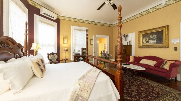 A four-poster bed with a canopy, a red sofa, and a small round table in a warmly lit room with a classic design.