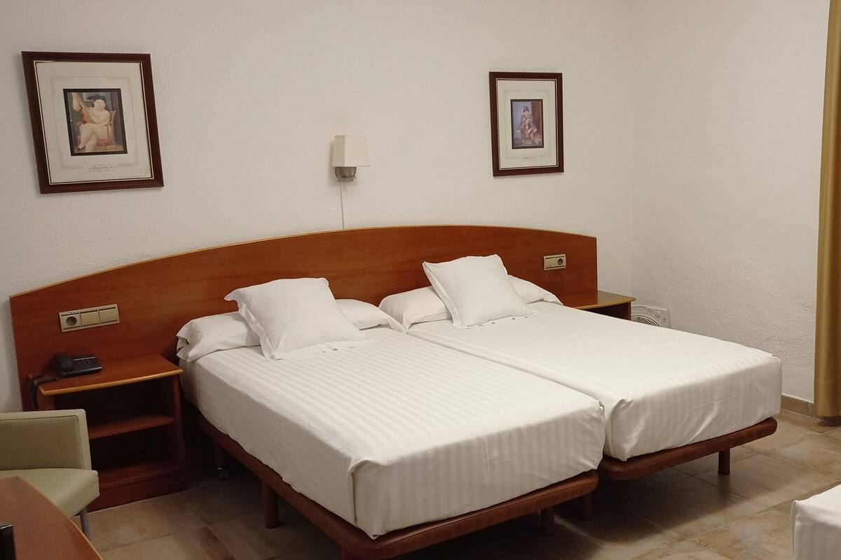 Habitación de hotel con dos camas individuales, una mesita de noche con lámpara y un escritorio pequeño con teléfono.